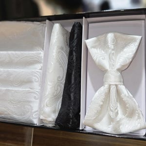 May include: A formal wear set in a black box. The set includes a white paisley bow tie, a black paisley ascot, and white paisley pocket squares. The bow tie is pre-tied. The set is designed for formal occasions.