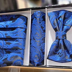 May include: A formal set featuring a black and blue paisley pattern. The set includes a folded pocket square, a necktie, and a bow tie, all neatly arranged in a box. The fabric has a subtle sheen, enhancing the intricate design.