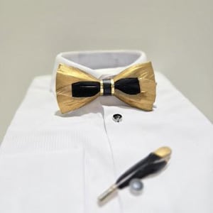 Puede incluir: Una pajarita dorada y negra y un alfiler de solapa a juego descansan sobre una camisa blanca. La pajarita tiene un centro negro y secciones exteriores doradas. El alfiler de solapa tiene una punta dorada.