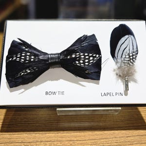 May include: A black feather bow tie with white polka dots and a black leather band. A matching feather lapel pin with a silver pin is also displayed. The words "BOW TIE" and "LAPEL PIN" are printed on the display.
