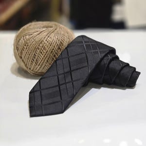 Peut inclure: Une cravate noire à motif géométrique est enroulée sur une surface blanche. Une pelote de ficelle est derrière la cravate. La cravate présente un motif texturé en forme de grille. L'arrière-plan est flou.