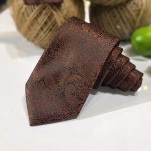 May include: A dark brown necktie featuring a detailed, intricate black paisley pattern woven into the fabric. The tie is folded in a layered, accordion-style arrangement, showcasing the textured surface and the deep, rich color contrast of the design.