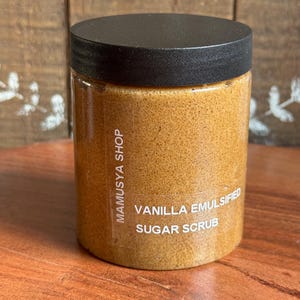 May include: A clear plastic jar with a black lid, filled with a light brown, textured sugar scrub. The label reads "MAMUSYA SHOP" and "VANILLA EMULSIFIED SUGAR SCRUB". The jar sits on a wooden surface.