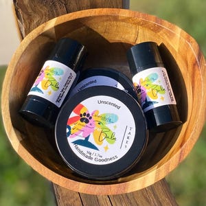Könnte beinhalten: Eine Holzschale enthält zwei schwarze Tuben und zwei schwarze Dosen mit Tierbalsam. Die Tuben und Dosen sind mit "Calendula Pet Balm" und "Unscented" beschriftet. Die Etiketten haben ein farbenfrohes Blumendesign.