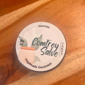 Könnte beinhalten: Eine runde, silberfarbene Dose Comfrey Salbe, beschriftet mit "Unscented" und "Handmade Goodness". Das Etikett zeigt eine pfirsichfarbene Illustration und Text. Die Dose enthält 28g Produkt.