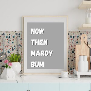 May include: Framed art print with the words "NOW THEN MARDY BUM" in white, bold, block letters on a gray background. The print is in a light wood frame, displayed in a kitchen setting with a light blue cabinet and decorative elements.