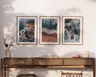 May include: Three framed photographs of ocean waves crashing against rocky cliffs. The photos are hung on a white wall above a wooden console table with a woven rattan top.