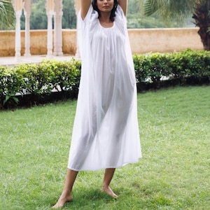 Puede incluir: Un vestido blanco de algodón con un escote fruncido y una silueta suelta y fluida. El vestido lo lleva una persona de pie en un campo de hierba.