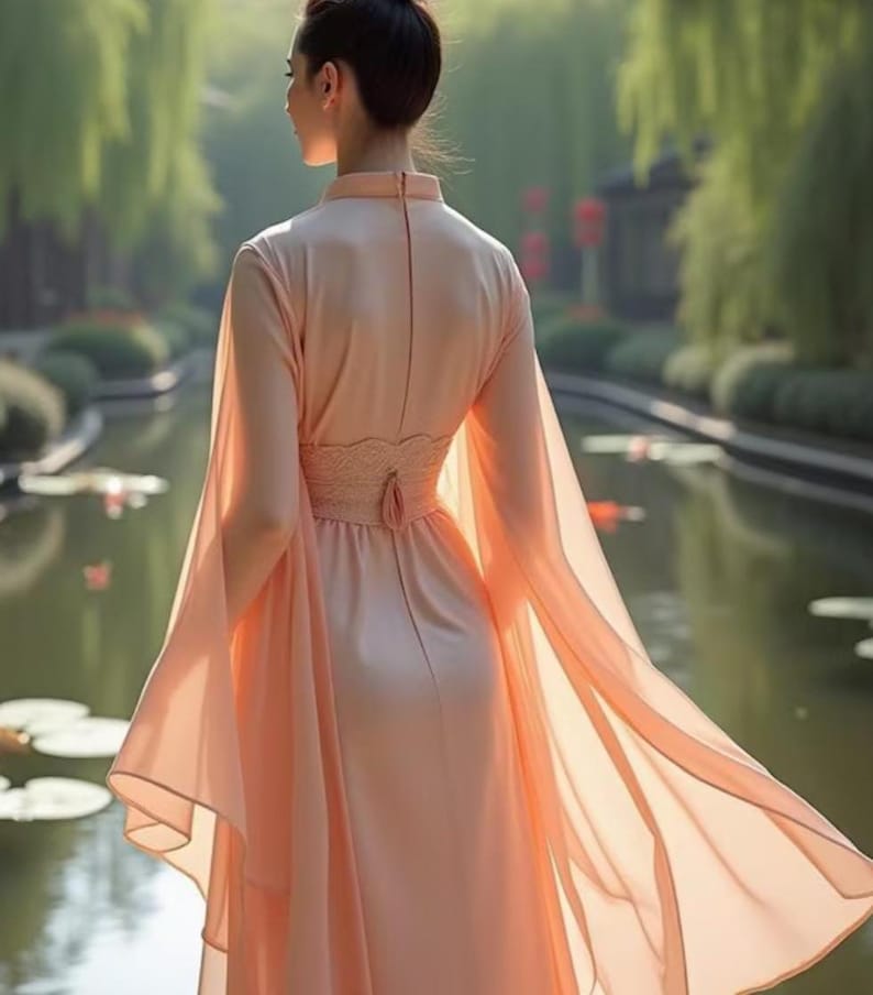 May include: A peach-colored dress with long, flowing sleeves and a high neckline. The dress features a fitted bodice with a lace detail at the waist and a flowing skirt. The model is standing near a body of water.