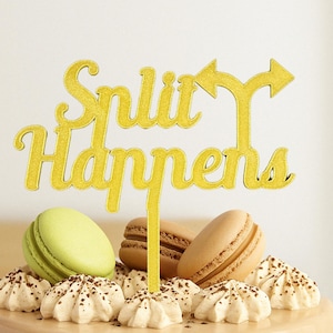 Könnte beinhalten: Goldener Glitzer-Kuchentopper mit dem Text "Split Happens" und einem Doppelpfeil. Der Topper befindet sich auf einem Kuchen, der mit Macarons und weißer Zuckerglasur verziert ist.