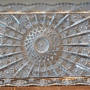 May include: A clear glass serving tray with a decorative, starburst pattern. The tray has a raised edge and a smooth, polished surface.