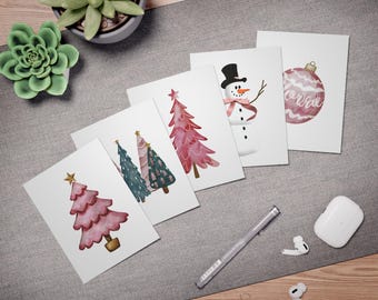 May include: A selection of Christmas cards with watercolour illustrations. Designs include pink Christmas trees, a snowman with a top hat and scarf, and a pink bauble. The cards are arranged on a grey surface with a succulent plant, pen, and earbuds.