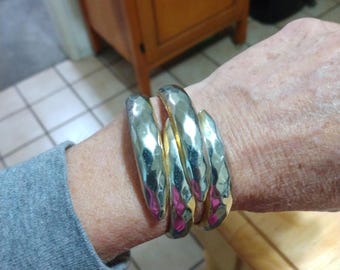May include: Four silver and gold-toned bangles with a hammered texture. The bracelets are stacked on a wrist, reflecting light and displaying a geometric design. The bangles are a statement piece of jewellery.