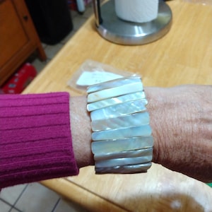 May include: A wide bracelet made of rectangular, iridescent mother-of-pearl pieces. The bracelet is worn on a wrist with a magenta sleeve visible. The bracelet's colors range from white to light gray.