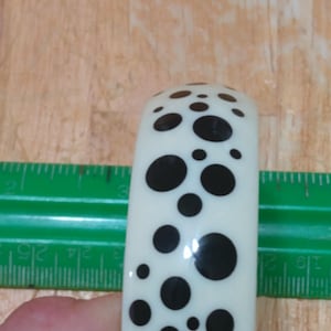 Puede incluir: Brazalete de color marfil con acabado brillante, adornado con un patrón de lunares negros de varios tamaños. El brazalete se muestra junto a una regla verde, para dar escala.