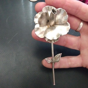 May include: A silver-toned metal rose brooch with detailed petals and a stem with two leaves. The brooch is held in a hand against a dark background. The flower is a detailed, three-dimensional design.