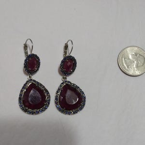 May include: A pair of dangle earrings with two red gemstone drops. The larger drop is surrounded by a silver setting with blue gemstones. The smaller drop is also surrounded by a silver setting with blue gemstones.