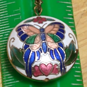 May include: A gold-toned necklace with a round pendant featuring a butterfly design. The butterfly has blue, green, and peach-colored wings, set against a white background. The pendant is accented with red and pink floral details.