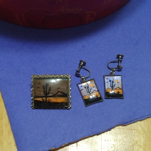 May include: A set of butterfly wing jewellery featuring a brooch and a pair of earrings. The jewellery is made with real butterfly wings and depicts a desert scene with a cactus and a setting sun. The brooch is silver-toned and has a square shape. The earrings are silver-toned and have a rectangular shape.