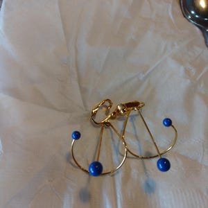 Puede incluir: Un par de pendientes de oro con cuentas azules. Los pendientes tienen forma de línea curva con un pequeño círculo en el extremo.
