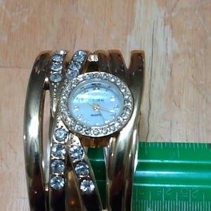 Könnte beinhalten: Goldfarbene Armbanduhr mit rundem Zifferblatt und Quarzwerk. Die Uhr hat ein helles Zifferblatt mit dem Wort "ORDEN" und ist mit klaren Kristallen verziert. Das Armband ist etwa 5 cm breit.