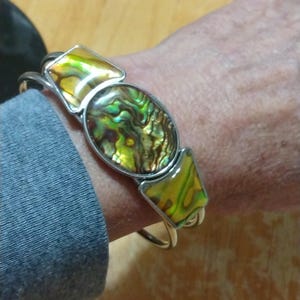 May include: Silver-toned cuff bracelet featuring three iridescent abalone shell inlays. The central inlay is oval, flanked by two diamond-shaped inlays. The abalone shells display a mix of green, gold, and brown hues, set in a silver metal frame.
