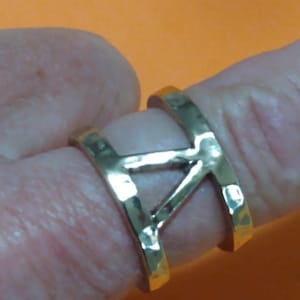 Puede incluir: Un anillo dorado con textura martillada. El anillo presenta un diseño estilizado de la letra "A", con una barra horizontal que conecta los elementos verticales. El anillo se lleva en un dedo sobre un fondo naranja.