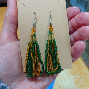 Peut inclure: Une paire de boucles d'oreilles à pompons perlées. Les boucles d'oreilles présentent des perles vertes et dorées, avec des crochets d'oreilles argentés. Les pompons sont fixés à une petite carte de présentation rectangulaire en carton.