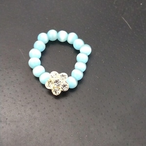 Puede incluir: Un anillo de cuentas azul claro con una pieza central en forma de flor. El anillo está hecho de pequeñas cuentas redondas de color azul claro. La pieza central en forma de flor es dorada y adornada con cristales transparentes.
