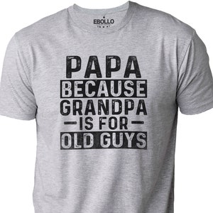 May include: A light gray t-shirt with the text "PAPA BECAUSE GRANDPA IS FOR OLD GUYS" in black, distressed lettering. The shirt has a crew neck and short sleeves. The brand logo is visible at the top.