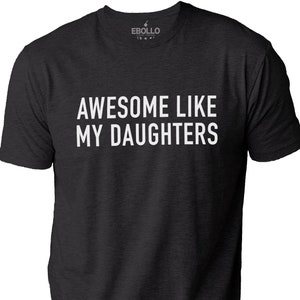 May include: Dark grey t-shirt featuring the white text "AWESOME LIKE MY DAUGHTERS". The shirt has a crew neck and short sleeves. The brand logo is visible at the top.