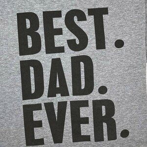 Puede incluir: Una camiseta gris con las palabras "BEST DAD EVER" impresas en letras negras grandes y audaces. El texto est&aacute; centrado y ocupa la mayor parte de la superficie de la camiseta. Dise&ntilde;o sencillo y cl&aacute;sico.