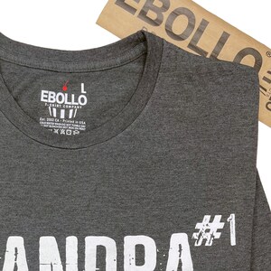 May include: A dark gray t-shirt with white text that reads "Grandpa #1". The shirt has a tag on the neck that reads "Ebollo T-Shirt Company" and includes care instructions.