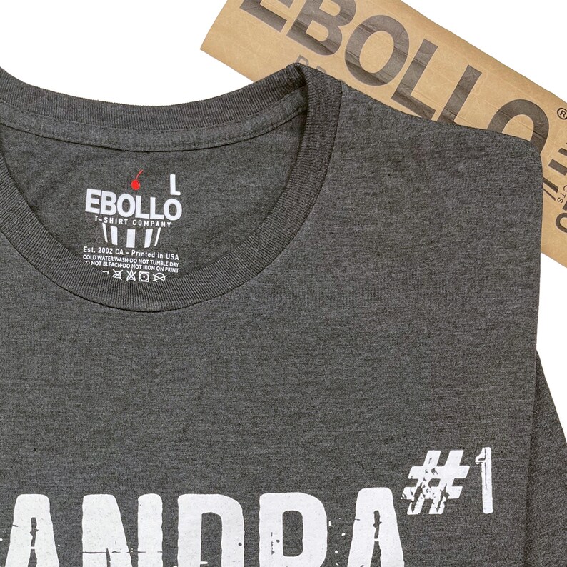 May include: A dark gray t-shirt with white text that reads "Grandpa #1". The shirt has a tag on the neck that reads "Ebollo T-Shirt Company" and includes care instructions.
