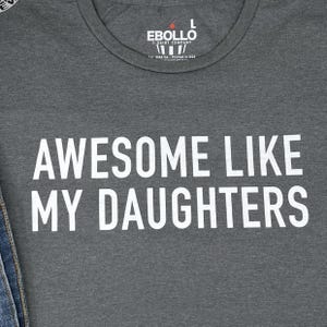 May include: A grey t-shirt featuring the white text "AWESOME LIKE MY DAUGHTERS". The shirt has a crew neck and the brand name "EBOLLO" is printed at the top. The t-shirt is made in the USA.