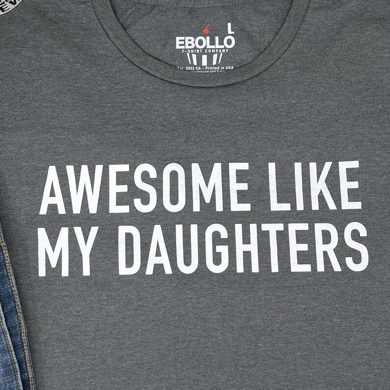 May include: A grey t-shirt featuring the white text "AWESOME LIKE MY DAUGHTERS". The shirt has a crew neck and the brand name "EBOLLO" is printed at the top. The t-shirt is made in the USA.