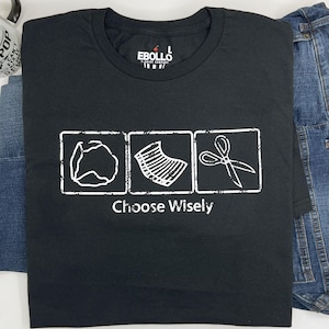 May include: Black t-shirt with a white graphic of three icons in squares. The icons are a crumpled piece of paper, a stack of papers, and a pair of scissors. The text "Choose Wisely" is below the icons.