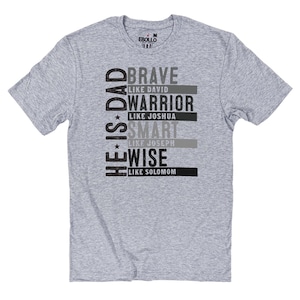 May include: A light gray t-shirt with the text "HE IS DAD" vertically on the left side. The shirt features the words "BRAVE," "WARRIOR," "SMART," and "WISE" with associated names, in a black and gray font.