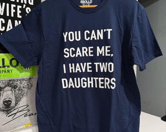 May include: A navy blue short-sleeved t-shirt with white text printed on the front that reads, "YOU CAN'T SCARE ME, I HAVE TWO DAUGHTERS." The shirt features a crew neckline and is displayed on a wooden hanger with an EBOLLO brand label.
