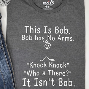Pu&ograve; includere: Maglietta grigio scuro XL con testo bianco: "This Is Bob. Bob has No Arms." Sotto, un omino stilizzato e il testo "Knock Knock" "Who's There?" "It Isn't Bob."