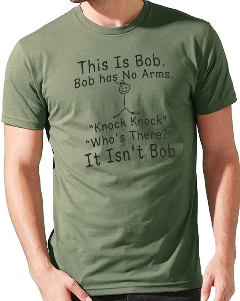 Pu&ograve; includere: T-shirt verde oliva con testo nero. Il testo recita: "This Is Bob. Bob has No Arms." Sotto c'&egrave; un omino stilizzato e il testo "Knock Knock" "Who's There?" "It Isn't Bob."