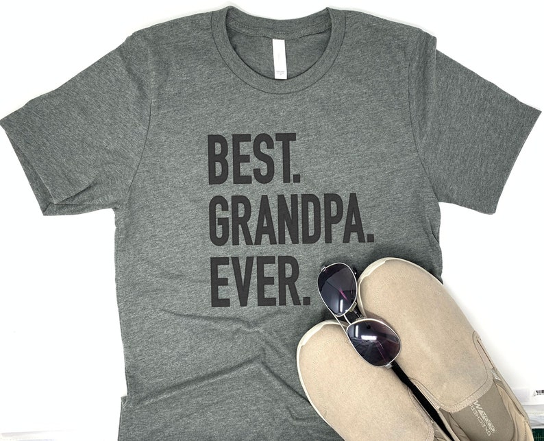 May include: A gray t-shirt with the text "BEST. GRANDPA. EVER." in black block letters. Sunglasses and beige slip-on shoes are placed on the shirt. The shirt has a crew neck and short sleeves.