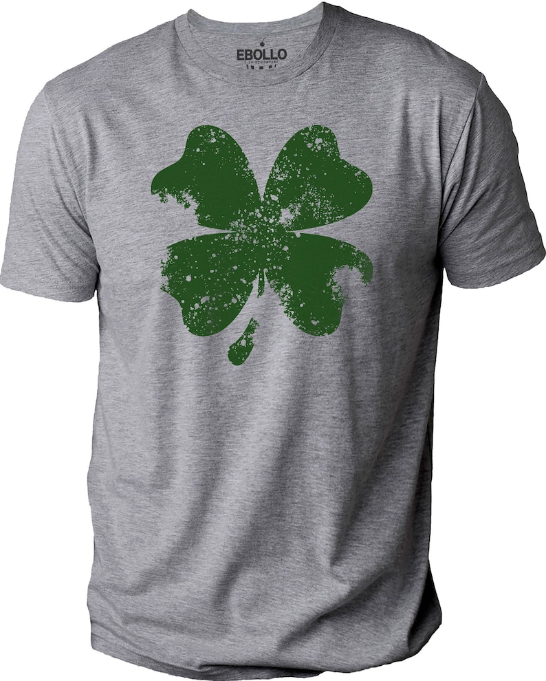 May include: A heather gray t-shirt featuring a distressed green four-leaf clover design. The shirt has short sleeves and a crew neck. The brand name "EBOLLO" is printed on the upper right of the shirt.