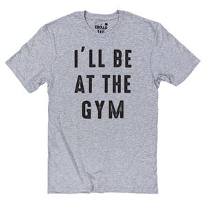 May include: A heather gray t-shirt with the text "I'LL BE AT THE GYM" in black, distressed lettering. The shirt has short sleeves and a crew neck. The brand name "EBULLO" is printed on the upper back of the shirt.