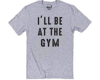 May include: A heather gray t-shirt with the text "I'LL BE AT THE GYM" in black, distressed lettering. The shirt has short sleeves and a crew neck. The brand name "EBULLO" is printed on the upper back of the shirt.