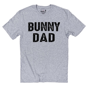 May include: A light gray t-shirt with the words "BUNNY DAD" printed in large, distressed black letters. The shirt has a crew neck and short sleeves. The brand name "EBULLO" is printed on the upper back of the shirt.