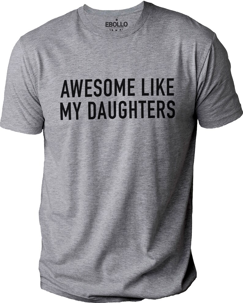 May include: A grey t-shirt featuring the black text "AWESOME LIKE MY DAUGHTERS". The shirt has a crew neck and short sleeves. The brand name "EBOLLO" is printed on the upper back of the shirt.
