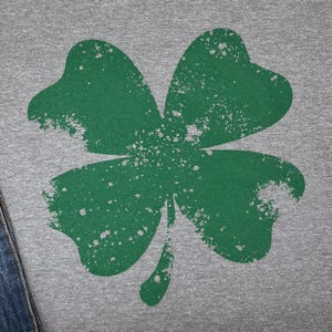 May include: A close-up of a gray t-shirt featuring a distressed green shamrock design. The four-leaf clover has a textured, worn appearance with white speckles. The shirt is partially visible, with a denim edge in the lower left corner.