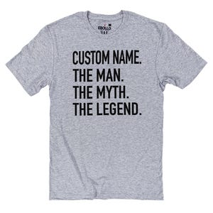 Op de afbeelding: Een lichtgrijs t-shirt met de tekst "CUSTOM NAME. THE MAN. THE MYTH. THE LEGEND." in vette zwarte letters. Het shirt heeft korte mouwen en een ronde hals.