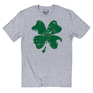 May include: A heather gray t-shirt featuring a large, distressed green shamrock design. The shamrock has a textured, worn appearance. The shirt has short sleeves and a crew neck. The brand name "EBOLLO" is printed on the collar.